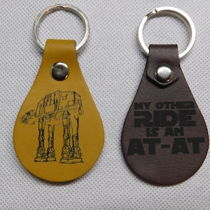 May include: Two leather keychains with metal key rings. One keychain is brown and features the text "My other ride is an AT-AT". The other keychain is yellow and features a black and white outline of an AT-AT.