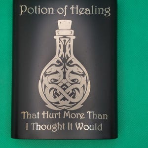 May include: A matte black metal flask featuring an engraved design of a potion bottle with intertwined serpent motifs. The text Potion of Healing is engraved above the bottle, and the text That Hurt More Than I Thought It Would is below.