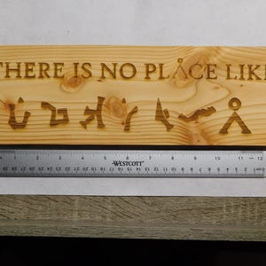 Puede incluir: Un letrero rectangular de madera con la frase "THERE IS NO PLACE LIKE" y símbolos tallados en la madera. Una regla metálica se coloca debajo del letrero. El letrero mide aproximadamente 30 cm de largo.