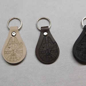 May include: Three leather keychains in beige, brown, and black, each featuring a detailed engraved design of a spaceship. The keychains have a teardrop shape and a silver key ring attached.