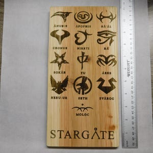 May include: A wooden plaque with the words "Stargate" and a series of symbols and names from the Stargate SG-1 television series. The symbols are in gold and the names are in black.