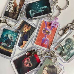 May include: A collection of rectangular acrylic keychains featuring various video game character and title art, including The Last of Us, Until Dawn, Resident Evil 2, and a purple rabbit character, all attached to silver metal split rings and clasps.