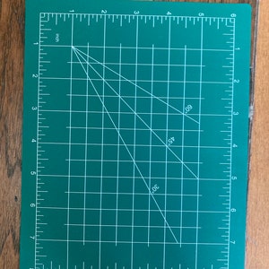 May include: A green self-healing cutting mat with white grid lines and markings. The mat has a ruler on all four sides, with markings in inches and degrees. The mat is marked with 30, 45, and 60 degrees.