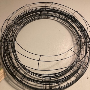 May include: A black metal wreath frame, made of multiple wire rings, is shown against a white wall. The frame is circular and has a diameter of approximately 12 inches.
