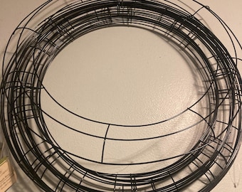 May include: A black metal wreath frame, made of multiple wire rings, is shown against a white wall. The frame is circular and has a diameter of approximately 12 inches.