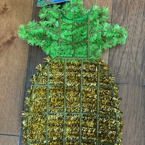May include: A gold and green pineapple-shaped Christmas ornament. The pineapple is made of tinsel and has a green top.