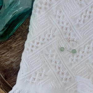May include: A pair of silver hoop earrings with small, light green beads. The earrings rest on a white, textured towel with a fringed edge. A teal glass bottle is in the background.