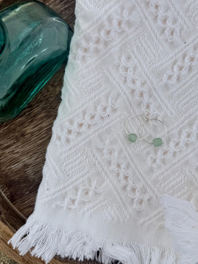 May include: A pair of silver hoop earrings with small, light green beads. The earrings rest on a white, textured towel with a fringed edge. A teal glass bottle is in the background.