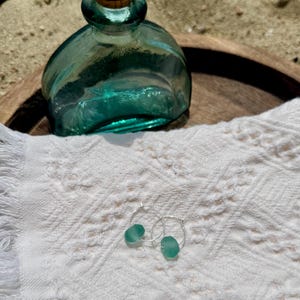 May include: A teal glass bottle with a cork stopper sits on a wooden tray next to a pair of silver hoop earrings, each with a teal sea glass bead. The items rest on a white, textured cloth.