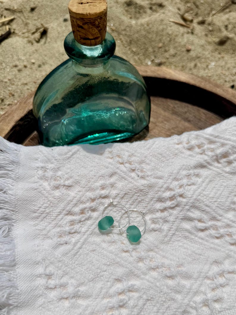 May include: A teal glass bottle with a cork stopper sits on a wooden tray next to a pair of silver hoop earrings, each with a teal sea glass bead. The items rest on a white, textured cloth.
