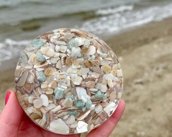 May include: A round coaster filled with small, colorful seashells and turquoise stones, encased in clear resin. The coaster is held in a hand, with a beach and ocean in the background. The coaster is a decorative item.
