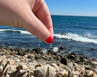 May include: A pair of silver hoop earrings, each adorned with a small, pale green, square-shaped stone. The earrings are held up against a backdrop of a bright blue sky and ocean, with a rocky shoreline in the foreground.
