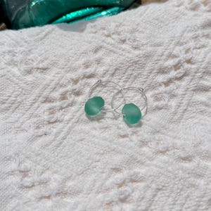 May include: A pair of silver hoop earrings, each with a rounded, seafoam green bead. The earrings rest on a white, textured surface, with a blurred turquoise glass object in the background. The earrings are a simple, elegant design.