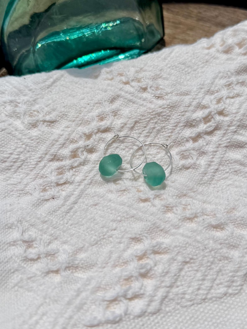 May include: A pair of silver hoop earrings, each with a rounded, seafoam green bead. The earrings rest on a white, textured surface, with a blurred turquoise glass object in the background. The earrings are a simple, elegant design.