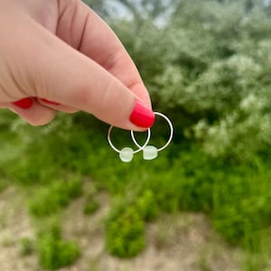 May include: A pair of silver hoop earrings, each with a small, pale green bead. The earrings are held up against a backdrop of green foliage and a cloudy sky. The earrings are a simple, minimalist design.