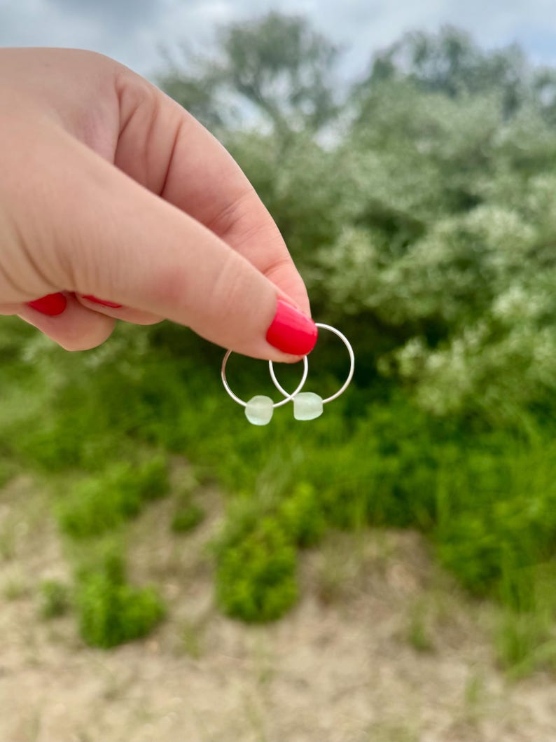 May include: A pair of silver hoop earrings, each with a small, pale green bead. The earrings are held up against a backdrop of green foliage and a cloudy sky. The earrings are a simple, minimalist design.
