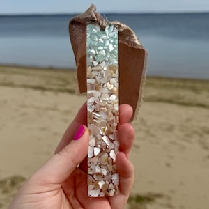 May include: A rectangular bookmark made of clear resin with embedded seashells. The bookmark is a gradient of light blue, white, and beige. The bookmark is held in a hand with pink nail polish.