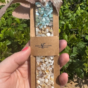 May include: A rectangular resin bookmark featuring embedded light blue and white stone fragments, attached to a taupe frayed ribbon. The bookmark is secured in a brown paper sleeve printed with the text Maeby One Day Design.