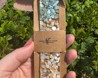 May include: A rectangular resin bookmark featuring embedded light blue and white stone fragments, attached to a taupe frayed ribbon. The bookmark is secured in a brown paper sleeve printed with the text Maeby One Day Design.