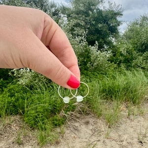 May include: A pair of silver hoop earrings with small, light green, square-shaped beads. The earrings are held up against a backdrop of green grass, trees, and a sandy beach.