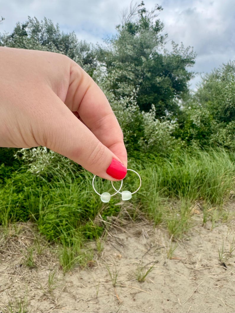 May include: A pair of silver hoop earrings with small, light green, square-shaped beads. The earrings are held up against a backdrop of green grass, trees, and a sandy beach.