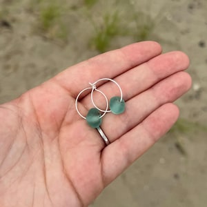 May include: A pair of silver hoop earrings, each adorned with a smooth, seafoam green glass bead. The earrings are displayed against a blurred background of sand and greenery, highlighting the natural beauty of the jewelry.