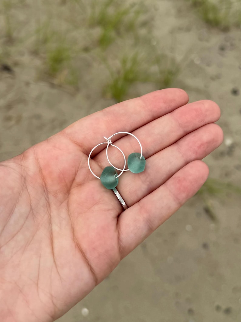 May include: A pair of silver hoop earrings, each adorned with a smooth, seafoam green glass bead. The earrings are displayed against a blurred background of sand and greenery, highlighting the natural beauty of the jewelry.