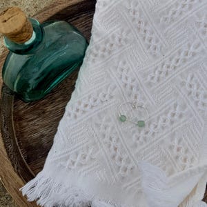 May include: A teal glass bottle with a cork stopper sits on a wooden tray next to a white fringed towel. A pair of silver hoop earrings with small, light green beads rests on the towel. The scene evokes a spa or beach theme.