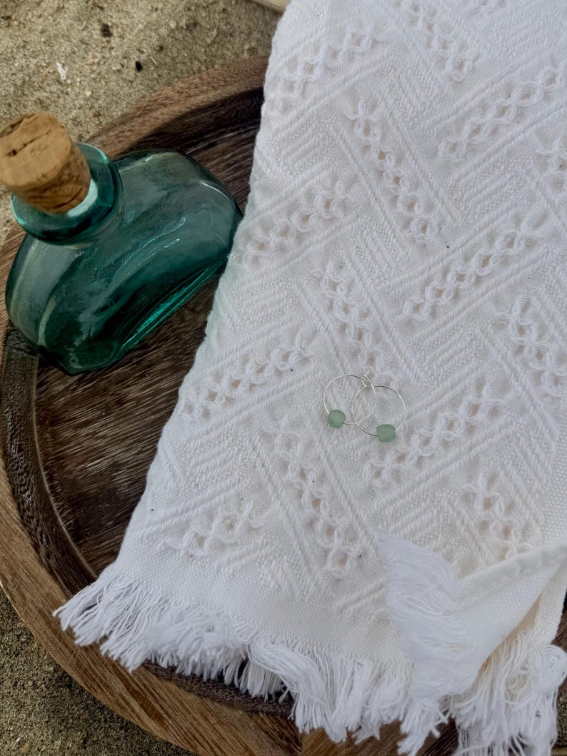 May include: A teal glass bottle with a cork stopper sits on a wooden tray next to a white fringed towel. A pair of silver hoop earrings with small, light green beads rests on the towel. The scene evokes a spa or beach theme.