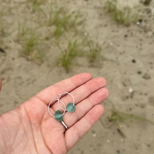 May include: A pair of silver hoop earrings, each adorned with a smooth, seafoam green, teardrop-shaped bead. The earrings are held in a hand against a backdrop of sand and beach grass, suggesting a coastal theme.
