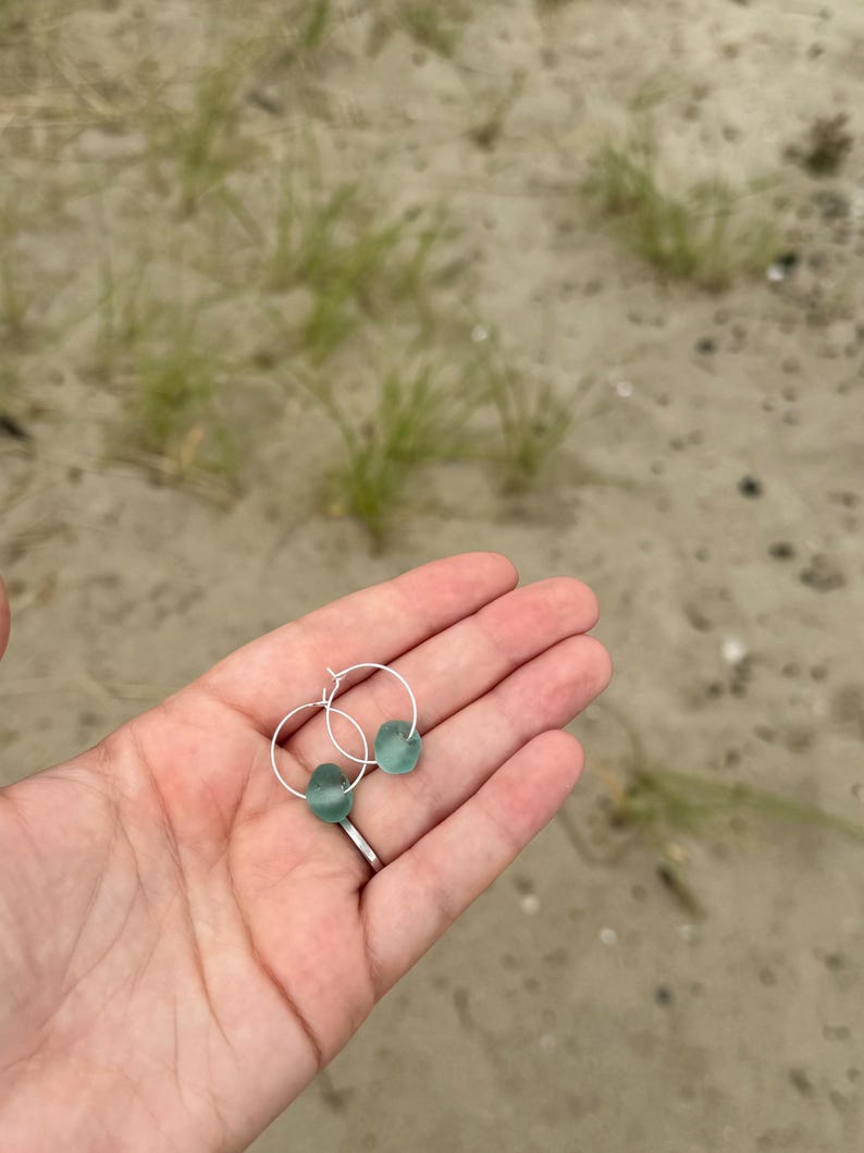 May include: A pair of silver hoop earrings, each adorned with a smooth, seafoam green, teardrop-shaped bead. The earrings are held in a hand against a backdrop of sand and beach grass, suggesting a coastal theme.