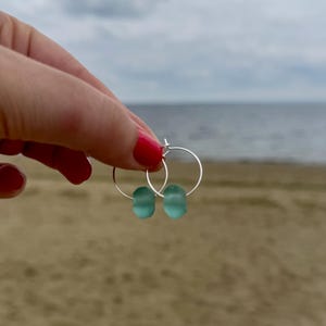 May include: Silver hoop earrings with two seafoam green, oval-shaped beads. The earrings are held up against a backdrop of a sandy beach and a cloudy sky. The earrings are a simple, elegant design.