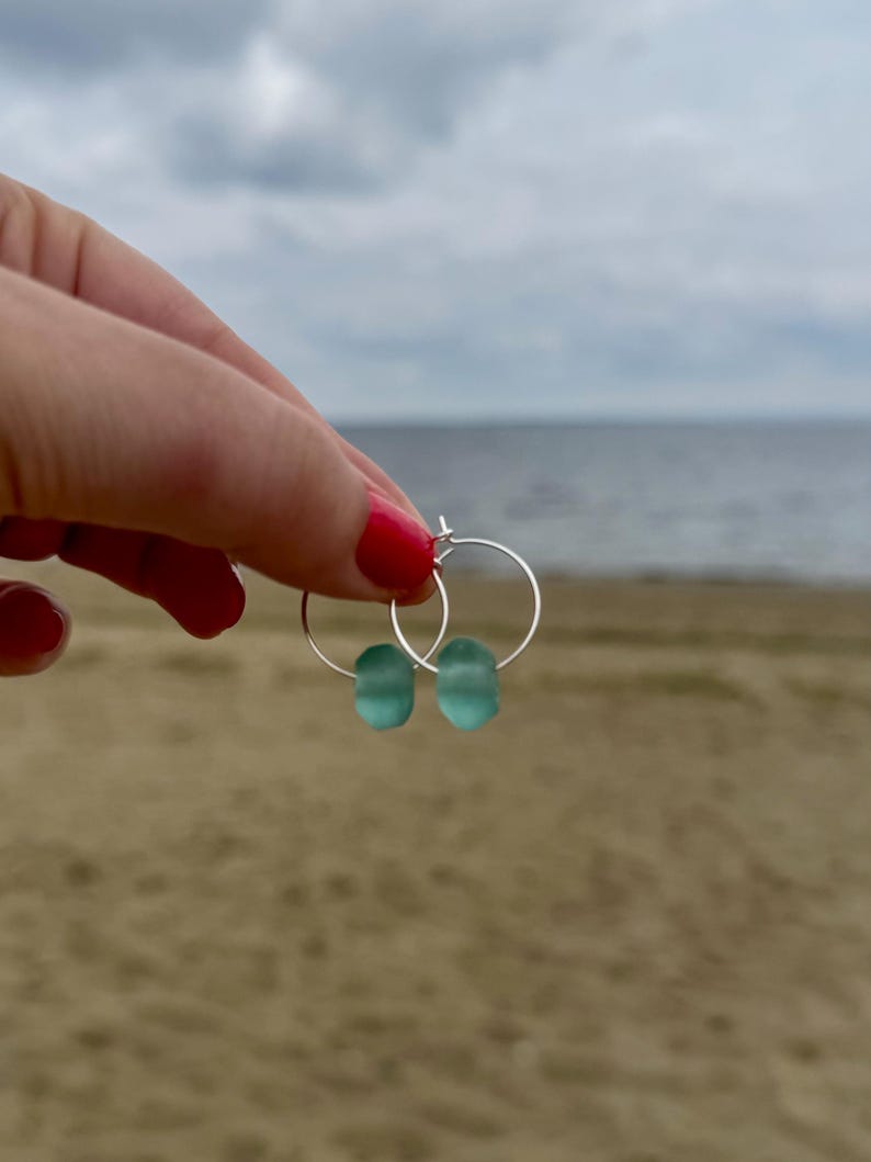 May include: Silver hoop earrings with two seafoam green, oval-shaped beads. The earrings are held up against a backdrop of a sandy beach and a cloudy sky. The earrings are a simple, elegant design.