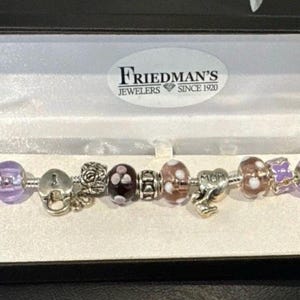 May include: A charm bracelet with various beads and charms, including purple glass beads, a heart-shaped lock, and a rose charm. The bracelet is displayed in a black jewelry box with the "Friedman's Jewelers Since 1920" logo.