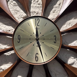 May include: A sunburst wall clock featuring a circular gold-toned face with black numerals at the 12, 3, 6, and 9 positions. The clock has black hands and a central starburst design, surrounded by long, curved, dark brown wooden rays.
