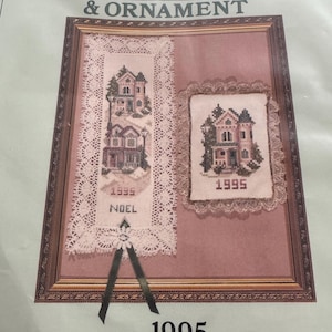 May include: A framed Christmas bookmark and ornament kit. The kit includes two cross-stitch designs of houses, one rectangular and one square, both with the year 1995. The rectangular design has the word "NOEL" stitched on it. The frame is brown.