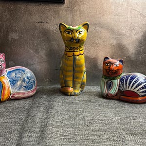 May include: Three hand-painted ceramic cat figurines. The left cat is pink and blue with floral patterns. The center cat is yellow with black stripes and sits upright. The right cat is orange and blue with a white fan-shaped pattern on its side.