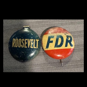 May include: Two circular political campaign buttons. The left button is dark blue with the yellow text ROOSEVELT. The right button features a red and yellow horizontal striped background with the blue text FDR and a metal pin attached at the bottom.