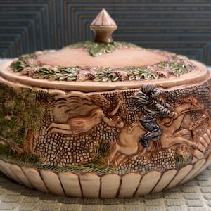 May include: A decorative ceramic lidded bowl with a detailed relief design. The bowl features a hunting scene with figures on horseback and a textured, patterned surface. The lid is topped with a finial and adorned with green leaf accents.