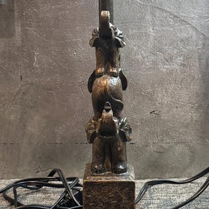 May include: Bronze-colored table lamp featuring a stacked elephant design. The lamp has a square base and a black lamp holder. The electrical cord is black. The lamp is a decorative home decor item.