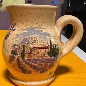 May include: A cream-colored ceramic pitcher with a speckled finish and a curved handle. The front features a detailed illustration of a stone house surrounded by lavender fields, tall cypress trees, and a large mountain under a blue sky.