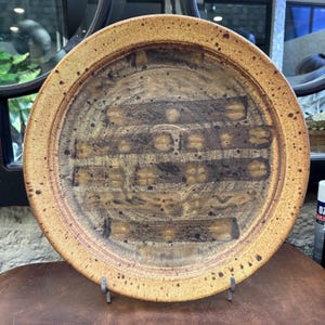 May include: A round, ceramic plate with a speckled, tan rim and a brown, textured center. The center features a series of horizontal and vertical rectangular shapes with a speckled design. The plate is displayed on a metal stand.
