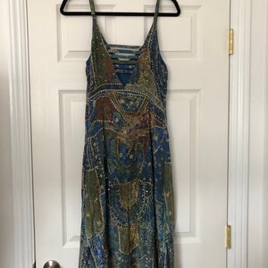 May include: A patterned dress in shades of blue, green, and brown, featuring spaghetti straps and a V-neck. The dress, adorned with sequins, hangs on a black hanger. It is a knee-length dress with a flowing design.