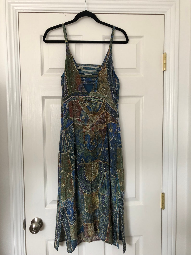 May include: A patterned dress in shades of blue, green, and brown, featuring spaghetti straps and a V-neck. The dress, adorned with sequins, hangs on a black hanger. It is a knee-length dress with a flowing design.