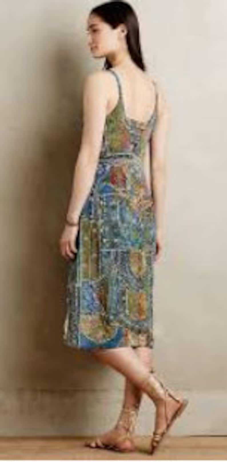 May include: A patterned midi dress in shades of blue, green, and gold. The dress has a sleeveless design and a fitted bodice that flows into a slightly flared skirt. The dress is worn with brown sandals. The pattern has a patchwork style.