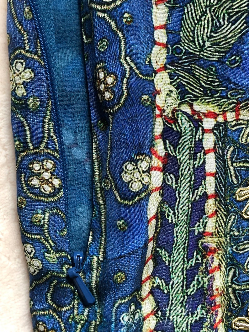 May include: Close-up of a blue patterned fabric with a zipper. The design features floral and geometric motifs in white, green, and red. The fabric has a contrasting blue lining, creating a rich, textured appearance.