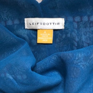 May include: A close-up shot of a blue fabric garment with a V-neck. The garment features a sheer overlay with a delicate pattern. The label reads "LEIFSDOTTIR" and a yellow tag states "MADE IN INDIA".