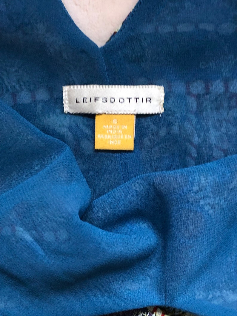 May include: A close-up shot of a blue fabric garment with a V-neck. The garment features a sheer overlay with a delicate pattern. The label reads "LEIFSDOTTIR" and a yellow tag states "MADE IN INDIA".