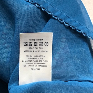 May include: Close-up of blue patterned fabric with a decorative edge. A white tag is visible, with text in English and Spanish, including "Dry Clean Only" and "Hecho en India". The fabric is likely part of a garment.