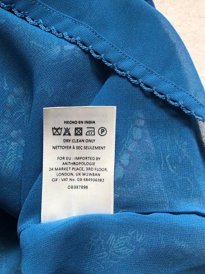 May include: Close-up of blue patterned fabric with a decorative edge. A white tag is visible, with text in English and Spanish, including "Dry Clean Only" and "Hecho en India". The fabric is likely part of a garment.